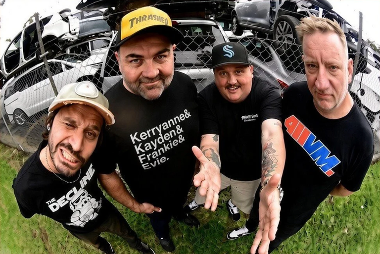 Punk rock band Not Dollar Short members posing outdoors in front of stacked cars at a scrapyard, captured with a fisheye wide-angle lens.
